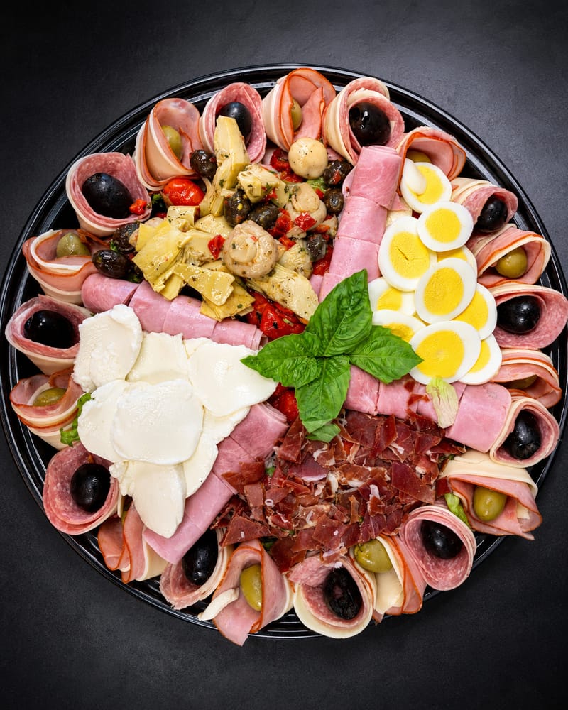 An antipasto platter on a black round tray, seen from above: rolled salami, ham and capicola with olives, fresh mozzarella, artichokes, marinated mushrooms, roasted peppers, sliced hard-boiled egg, prosciutto and basil