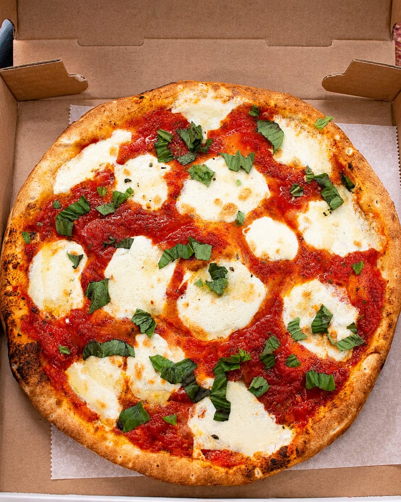 A margherita pizza in an open cardboard box, seen from above: red sauce, rounds of melted fresh mozzarella and torn basil on a puffed, blistered crust